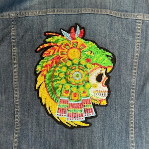 Puede incluir: Un parche colorido y con lentejuelas que presenta una calavera con un elaborado tocado. El diseño incorpora lentejuelas verdes, amarillas, rojas y blancas, y está adherido a una chaqueta vaquera. El parche tiene un borde negro.