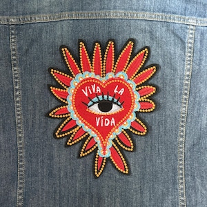 Può includere: Un cuore rosso ricamato con un occhio nero e le parole "Viva La Vida" su una giacca di jeans.