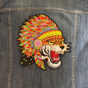 May include: A blue denim jacket featuring a large embroidered patch on the back. The patch depicts a tiger head wearing a vibrant, multi-colored feathered headdress with red, yellow, green, and orange tones, with the tiger's mouth open in a roar.