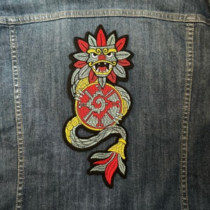 May include: A gray and red embroidered patch of a mythical creature holding a circular symbol. The creature has a large head with a beak and a long tail.