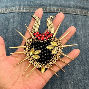 Peut inclure: Un cœur noir perlé avec des strass dorés et rouges, entouré de pointes dorées.