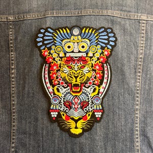 Puede incluir: Chaqueta vaquera con un parche bordado grande y colorido. El parche presenta un diseño de tigre estilizado con detalles amarillos, rojos, azules y blancos. La chaqueta es de mezclilla de lavado medio.