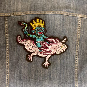 Puede incluir: Parche bordado con una figura turquesa con una corona dorada montada en un ajolote rosa. La figura sostiene una serpiente y una copa. El parche está cosido en una chaqueta vaquera.