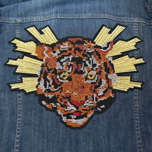 May include: A brown and orange embroidered tiger head patch with a yellow sunburst design. The patch is sewn onto a blue denim jacket.