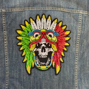 May include: A denim jacket with a colourful, sequined patch depicting a skull wearing a feathered headdress. The patch uses red, green, yellow, and white sequins, creating a vibrant design.