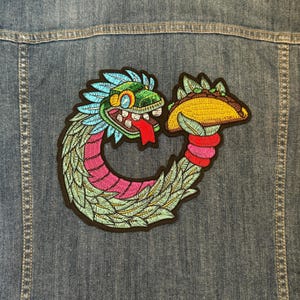 May include: An embroidered patch featuring a green, blue and pink serpent with feathers, eating a yellow taco. The serpent is curled into a circle and has a red tongue sticking out.