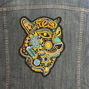 May include: A colorful embroidered patch of a jaguar's head with intricate floral and geometric designs in shades of blue, yellow, and orange. The patch is on a denim background.