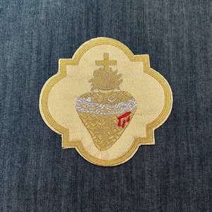 May include: A gold embroidered patch with a heart, thorns, and a cross. The heart is surrounded by a white rope and has a red drop of blood on it.