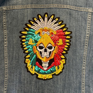 May include: A denim jacket with a large, colourful patch featuring a skull wearing a headdress. The patch is embellished with sequins in red, green, yellow, and white. The skull has a detailed design, and the jacket is a medium wash.