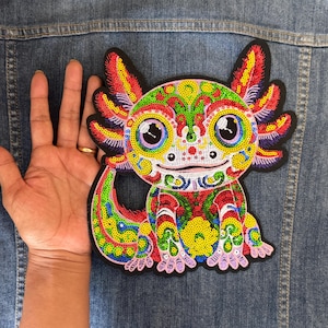 Puede incluir: Parche de axolotl colorido con un diseño detallado de lentejuelas. El parche presenta una vibrante mezcla de colores que incluyen verde, amarillo, rojo y azul. El axolotl tiene ojos grandes y expresivos y una boca sonriente. El parche mide aproximadamente 10 cm de ancho.