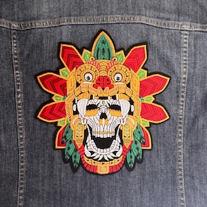 May include: A colourful Aztec skull design embroidered patch, with red, yellow, green, and white details, attached to a denim jacket. The patch showcases intricate artwork and vibrant colours. This patch is ideal for adding a unique touch to apparel.