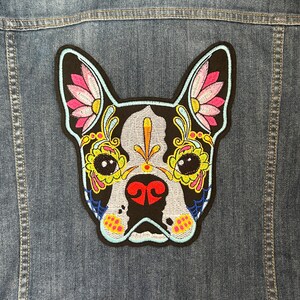 May include: Embroidered iron-on patch of a black and white Boston Terrier with colorful sugar skull details. The dog's eyes are outlined in yellow and have black pupils. The nose is red and the mouth is black. The ears are pink and white with floral designs.