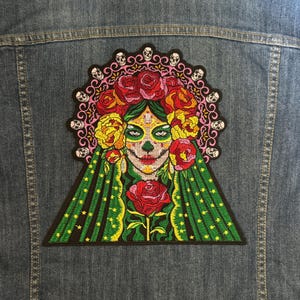 Puede incluir: Parche bordado para planchar con una mujer calavera de azúcar del Día de Muertos con una corona floral y un vestido verde y dorado. El parche tiene una combinación de colores rosa, verde, amarillo y blanco.
