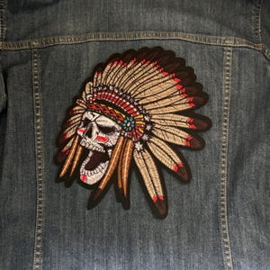 May include: Embroidered patch featuring a Native American headdress with a skull face. The patch has tan, red, and brown colors, with intricate feather details. It is attached to a denim jacket.