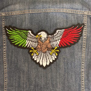 Puede incluir: Parche bordado de águila con alas verdes, blancas y rojas, que se asemeja a un diseño de bandera. El parche está cosido en una chaqueta vaquera. El águila tiene un cuerpo marrón, cabeza blanca y garras amarillas.