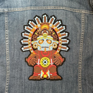 May include: An embroidered patch featuring a stylized, cartoon-like figure with a skull face, wearing a headdress with a sunburst design. The figure is brown, orange, yellow, and white.
