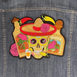 Puede incluir: Parche bordado con una calavera de azúcar colorida con sombrero, rodeada de botellas de tequila, rosas y chiles. El parche está adherido a una chaqueta vaquera, mostrando un tema mexicano festivo.