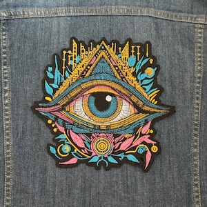 May include: Embroidered patch featuring a colorful eye within a triangle, set against a denim jacket. The patch has a detailed design with blue, yellow, pink, and black elements. The eye is the focal point, surrounded by floral and architectural motifs.