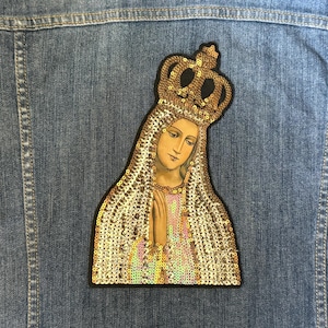 May include: A gold and white sequined patch depicting a woman wearing a crown and a white robe with her hands clasped in prayer.