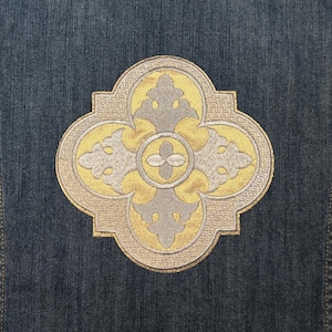 Puede incluir: Un diseño bordado en oro y plata sobre un fondo de mezclilla. El diseño es un trébol de cuatro hojas con un círculo central y detalles intrincados.