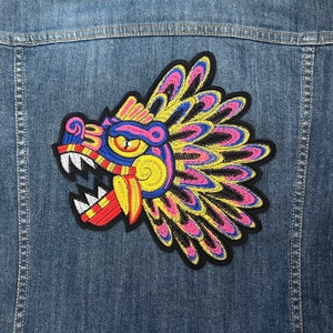 Può includere: Un ricamo colorato a forma di creatura mitologica con un copricapo piumato. La creatura ha una grande bocca aperta con denti affilati e un corpo con motivi gialli, rossi e blu.