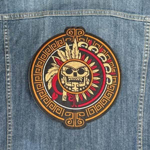 May include: A circular embroidered patch with a gold skull wearing a headdress with red and white accents. The skull is surrounded by a black and white geometric pattern.