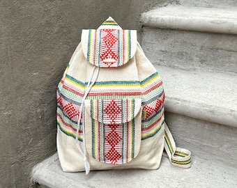 May include: Cream-colored backpack with a woven design featuring red, green, yellow, and white stripes. The backpack has a flap closure with a wooden toggle, a front pocket, and adjustable shoulder straps.