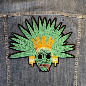 May include: Embroidered patch depicting an Aztec skull with a green face, large eyes, and a red mouth. The skull is decorated with a green headdress and gold details, set against a black border. The patch is on a denim jacket.