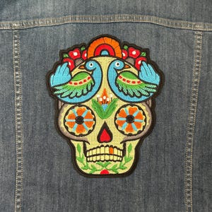 May include: Embroidered skull patch with a colorful design featuring two birds, flowers, and intricate details. The patch is attached to a denim jacket. The skull has orange, blue, and green accents.