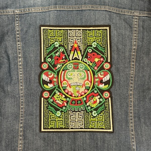 Peut inclure: Un patch brodé vert, noir et rouge avec un visage stylisé au centre. Le visage a de grands yeux et une large bouche. Le patch est entouré d'un motif géométrique.