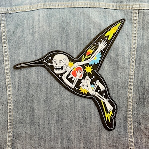 Könnte beinhalten: Schwarzer gestickter Patch mit einem Skelett-Kolibri mit bunten Blumen und Sternen. Der Kolibri fliegt mit ausgebreiteten Flügeln.