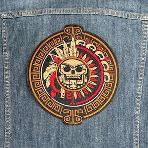 May include: A circular embroidered patch with a gold and red design. The center of the patch features a skull with a feathered headdress. The skull is surrounded by a circle of geometric patterns.