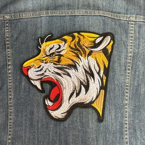 以下が含まれることがあります： 黄色と白の刺繍が施された虎の頭のワッペンで、赤い口が開いています。ワッペンは青いデニムジャケットに付いています。