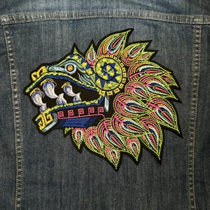 May include: A colorful embroidered patch depicting a mythical creature with a large head, sharp teeth, and a feathered headdress. The patch is on a denim background.