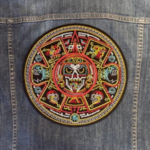 May include: Embroidered patch featuring a detailed Aztec calendar design with a central skull motif. The patch is circular, with vibrant colors including red, yellow, turquoise, and black. It is attached to a denim jacket.