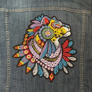 Puede incluir: Un parche bordado colorido que presenta una cabeza de animal estilizada con plumas y patrones geométricos. El animal es un guepardo o un leopardo con manchas amarillas y negras. El parche está sobre un fondo de mezclilla azul.