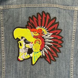 Pode incluir: Casaco de ganga com um grande e colorido patch com uma caveira com cabeça de águia e um cocar de penas. O patch é predominantemente amarelo, vermelho e preto, com detalhes intrincados.