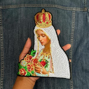 May include: A white and gold sequined patch of Our Lady of Fatima, a religious figure wearing a crown and holding a bouquet of red roses.
