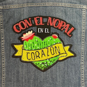 May include: A denim jacket with a colorful embroidered patch that says "CON EL NOPAL EN EL CORAZON". The patch features a green cactus heart with red flowers and yellow accents.