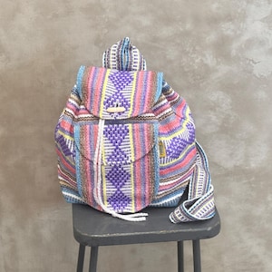 May include: A colorful woven backpack with a flap closure and a drawstring. The backpack features a pattern of vertical stripes in pink, purple, blue, and yellow. It has a wooden toggle closure and a small front pocket.