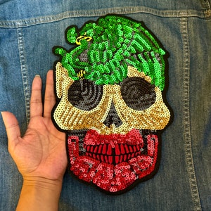 Op de afbeelding: Een pailletten schedel patch met een groen en zwart ontwerp. De schedel heeft een rode mond en een groen en zwart ontwerp bovenop het hoofd.