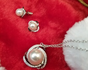 May include: A pearl jewelry set on a red background. The set includes a necklace with a large pearl pendant, a pair of stud earrings, and a pair of pearl earrings. The jewelry is silver-toned and has a halo of small, clear stones around the pearls.