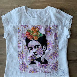 以下が含まれることがあります： 花柄のプリントが施された白いリネン製のTシャツ。黒と白のフリーダ・カーロの画像が描かれており、髪には花が飾られています。