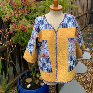 Puede incluir: Una chaqueta acolchada con estampado floral azul y blanco en la parte delantera y una espalda de color amarillo mostaza sólido. La chaqueta tiene cuello en V y mangas tres cuartos.