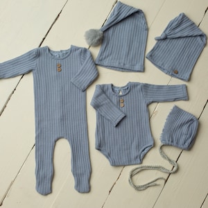 May include: A collection of baby clothes in a pale blue colour, featuring a footed all-in-one, a long-sleeved bodysuit, and three different style hats. Each item has a ribbed texture and wooden button details, ideal for newborn photos.