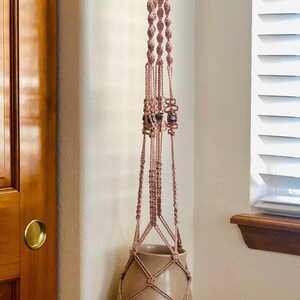 Peut inclure: Un porte-plante en macramé avec un pot en céramique brun. Le porte-plante est fait d'un cordon rose clair et a des perles en bois.