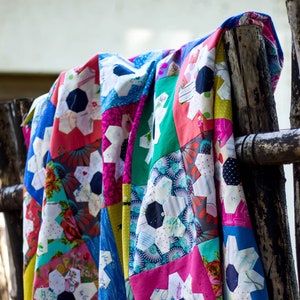 Pode incluir: Uma colcha de retalhos colorida com um design floral. A colcha é feita de vários tecidos em tons de rosa, azul, verde e branco. A colcha está pendurada em uma cerca de madeira.
