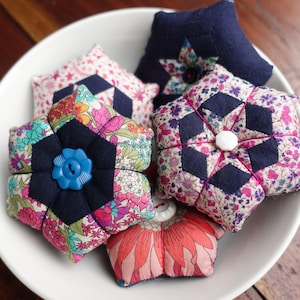 Może przedstawiać: Pięć kolorowych poduszek na szpilki w kształcie kwiatów, gwiazd i wzorów geometrycznych. Poduszki znajdują się w białej misce na drewnianej powierzchni.