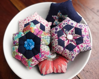May include: Five colorful fabric pincushions in the shape of flowers, stars, and geometric designs. The pincushions are in a white bowl on a wooden surface.