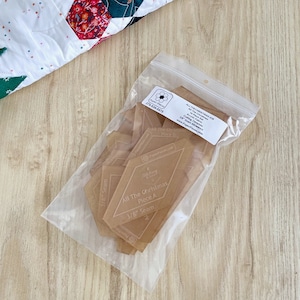 Peut inclure: Un sac en plastique transparent rempli de gabarits en plastique brun étiquetés "All The Christmas Piece A 3/8" Seam". Le sac est sur une surface en bois brun clair.